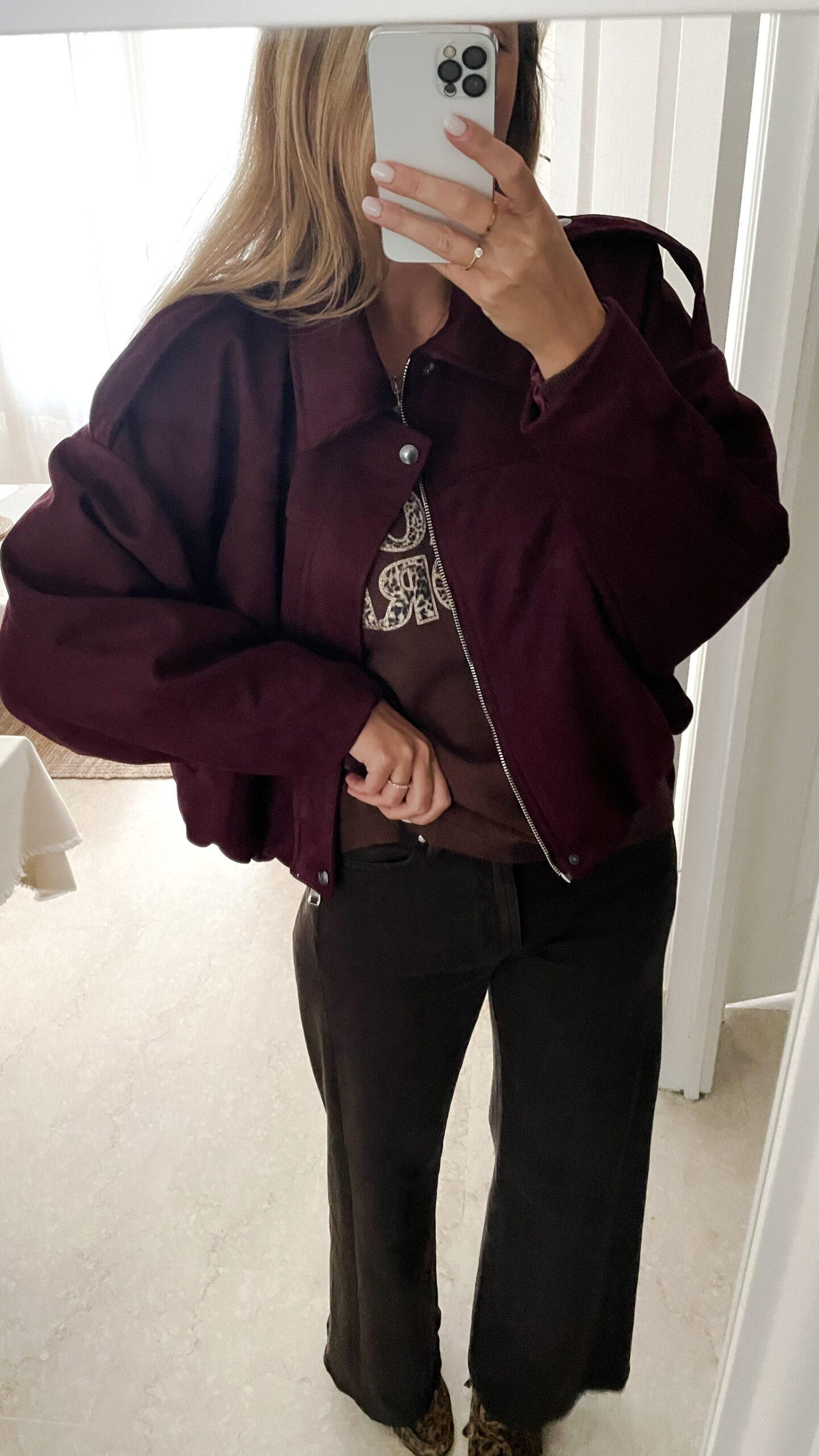 Femme portant une veste effet daim couleur bordeaux, assortie à un pull ton sur ton, une jupe noire et des collants foncés, dans un intérieur lumineux.