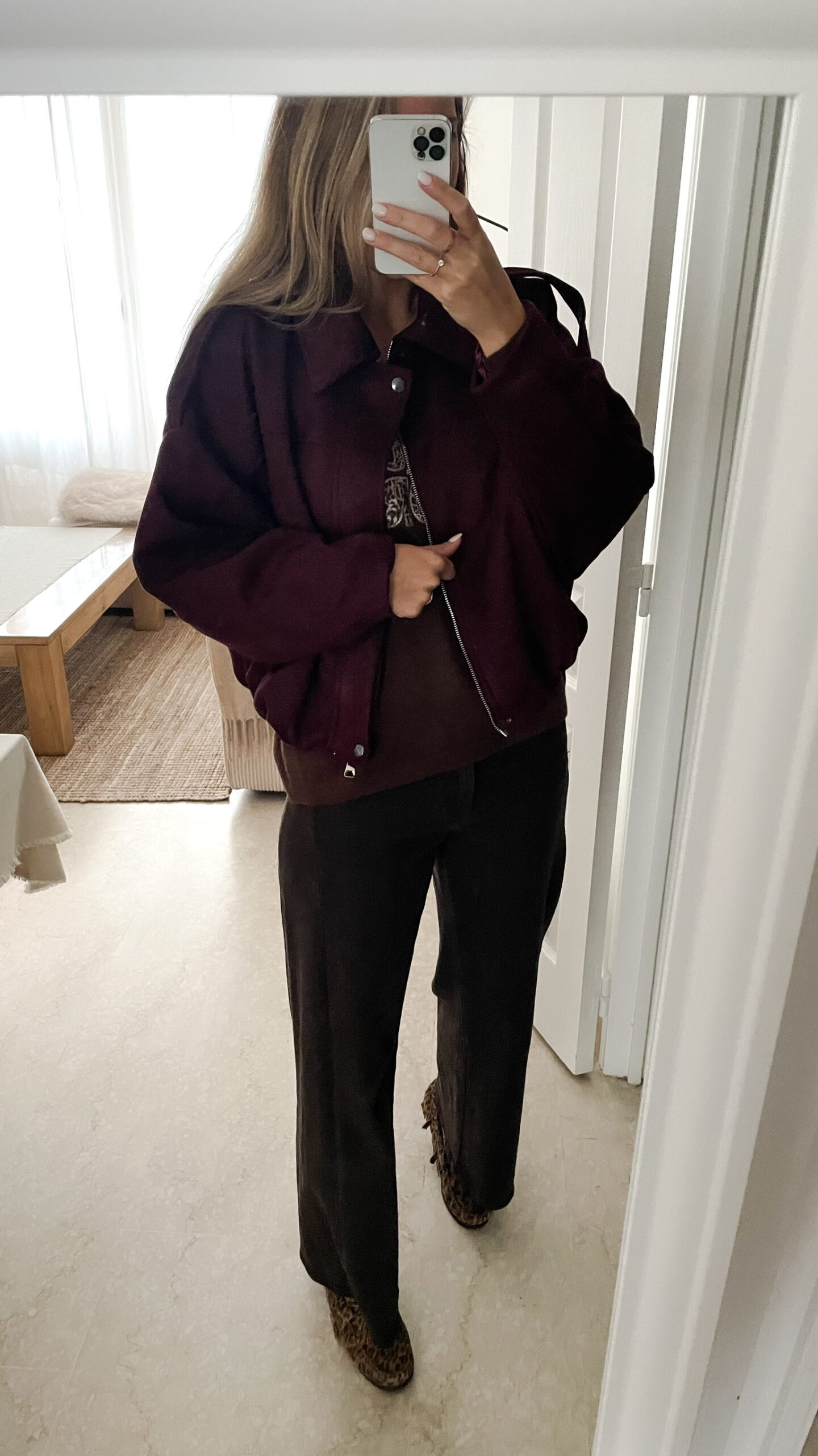 Femme portant une veste effet daim couleur bordeaux, assortie à un pull ton sur ton, une jupe noire et des collants foncés, dans un intérieur lumineux.