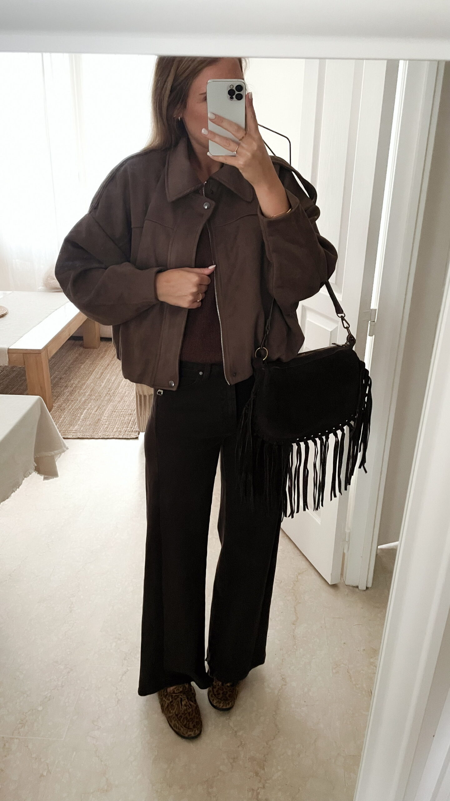 Femme portant une veste effet daim couleur marron taupe, assortie à un pull en alpaga taupe ton sur ton, un pantalon marron, une echarpe à motifs et un sac en daim marron avec des franges dans un intérieur lumineux.