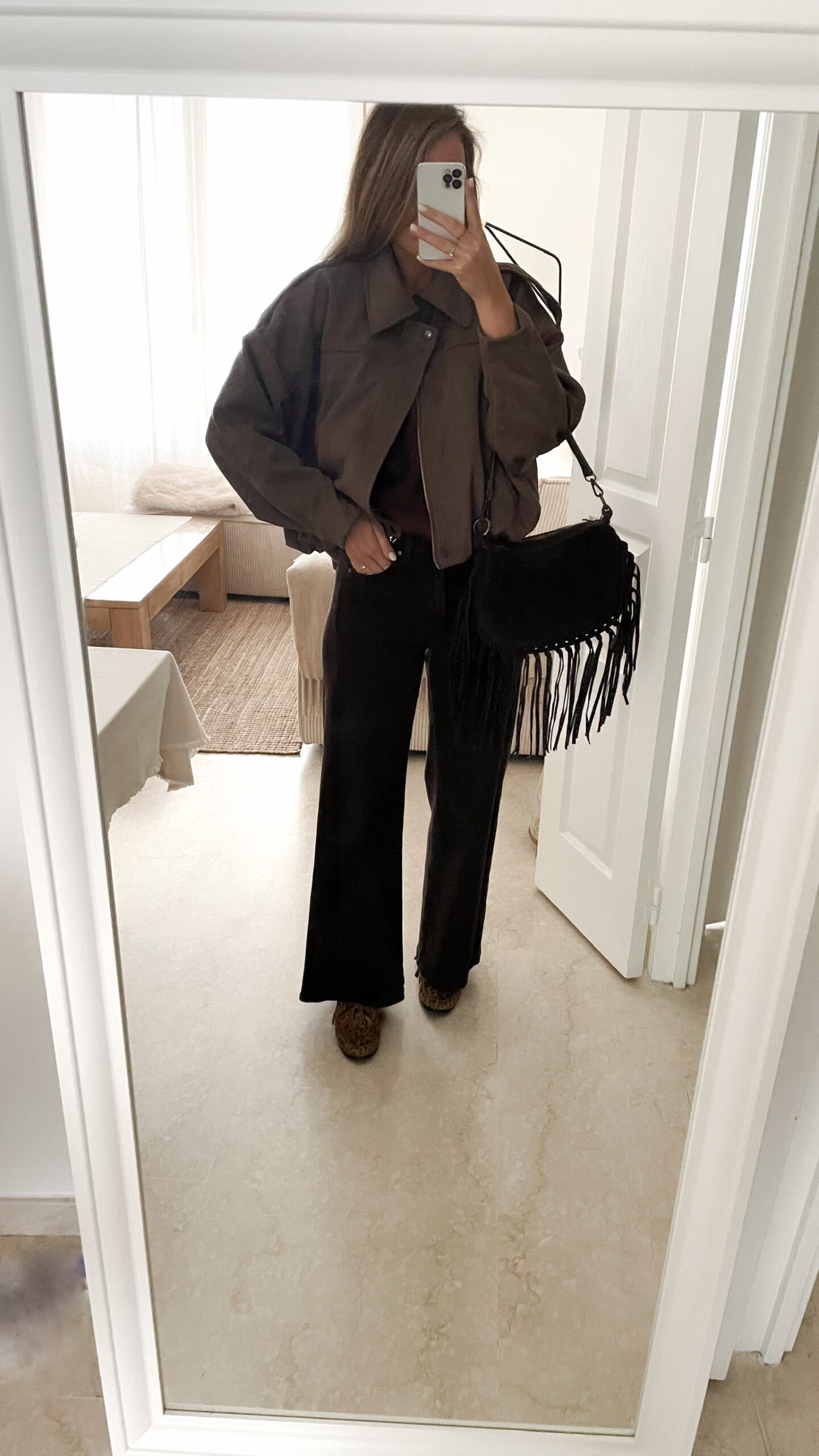 Femme portant une veste effet daim couleur marron taupe, assortie à un pull en alpaga taupe ton sur ton, un pantalon marron, une echarpe à motifs et un sac en daim marron avec des franges dans un intérieur lumineux.