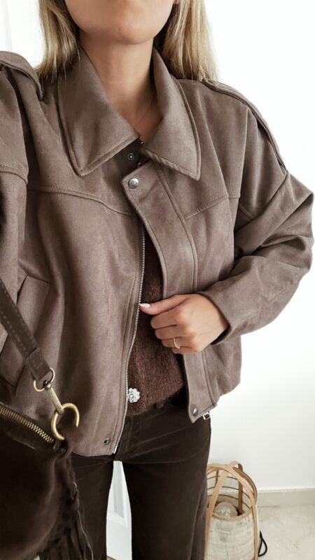 Femme portant une veste effet daim couleur marron taupe, assortie à un pull en alpaga taupe ton sur ton, un pantalon marron, une echarpe à motifs et un sac en daim marron avec des franges dans un intérieur lumineux.