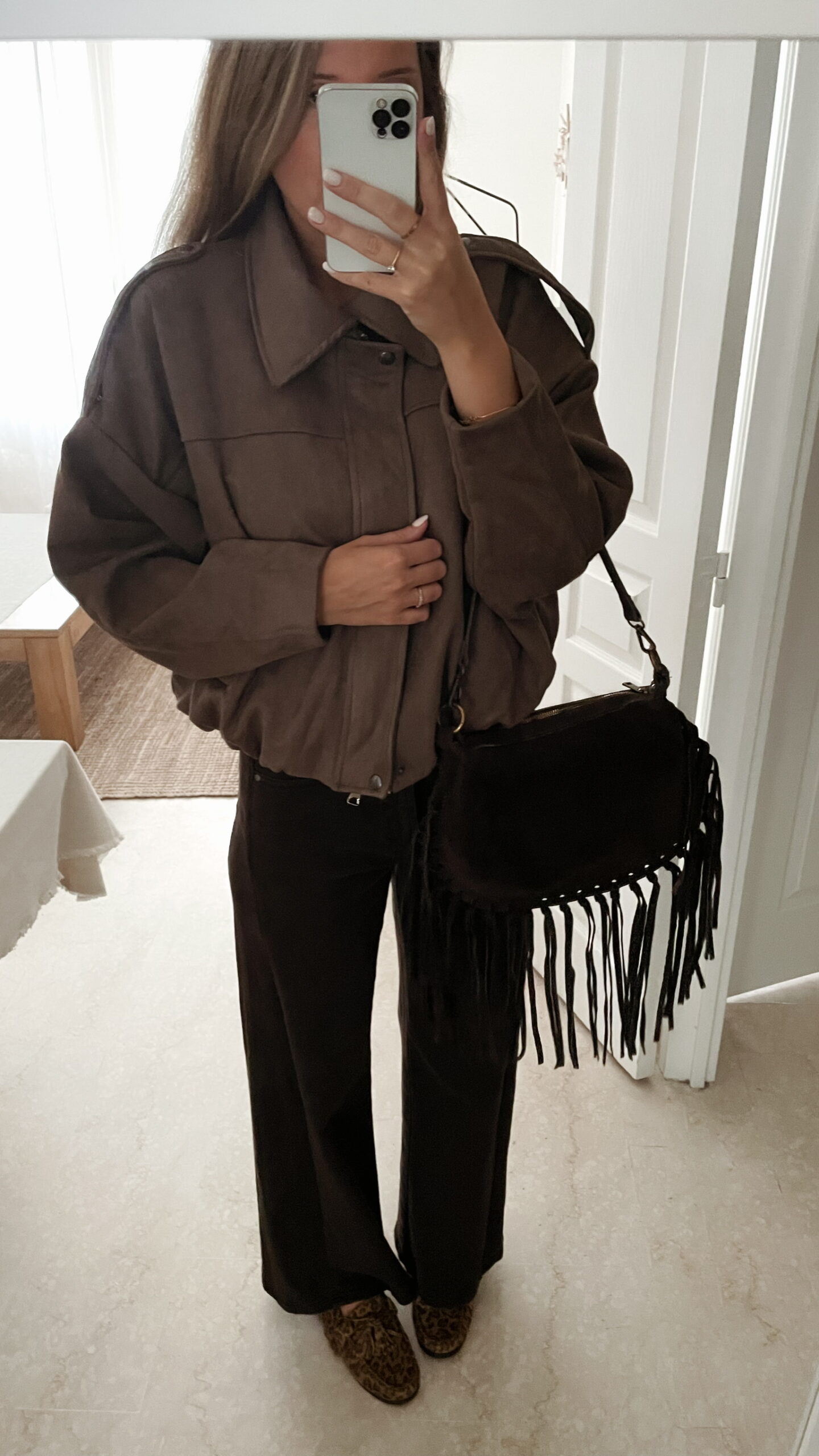 Femme portant une veste effet daim couleur marron taupe, assortie à un pull en alpaga taupe ton sur ton, un pantalon marron, et un sac en daim marron avec des franges dans un intérieur lumineux.