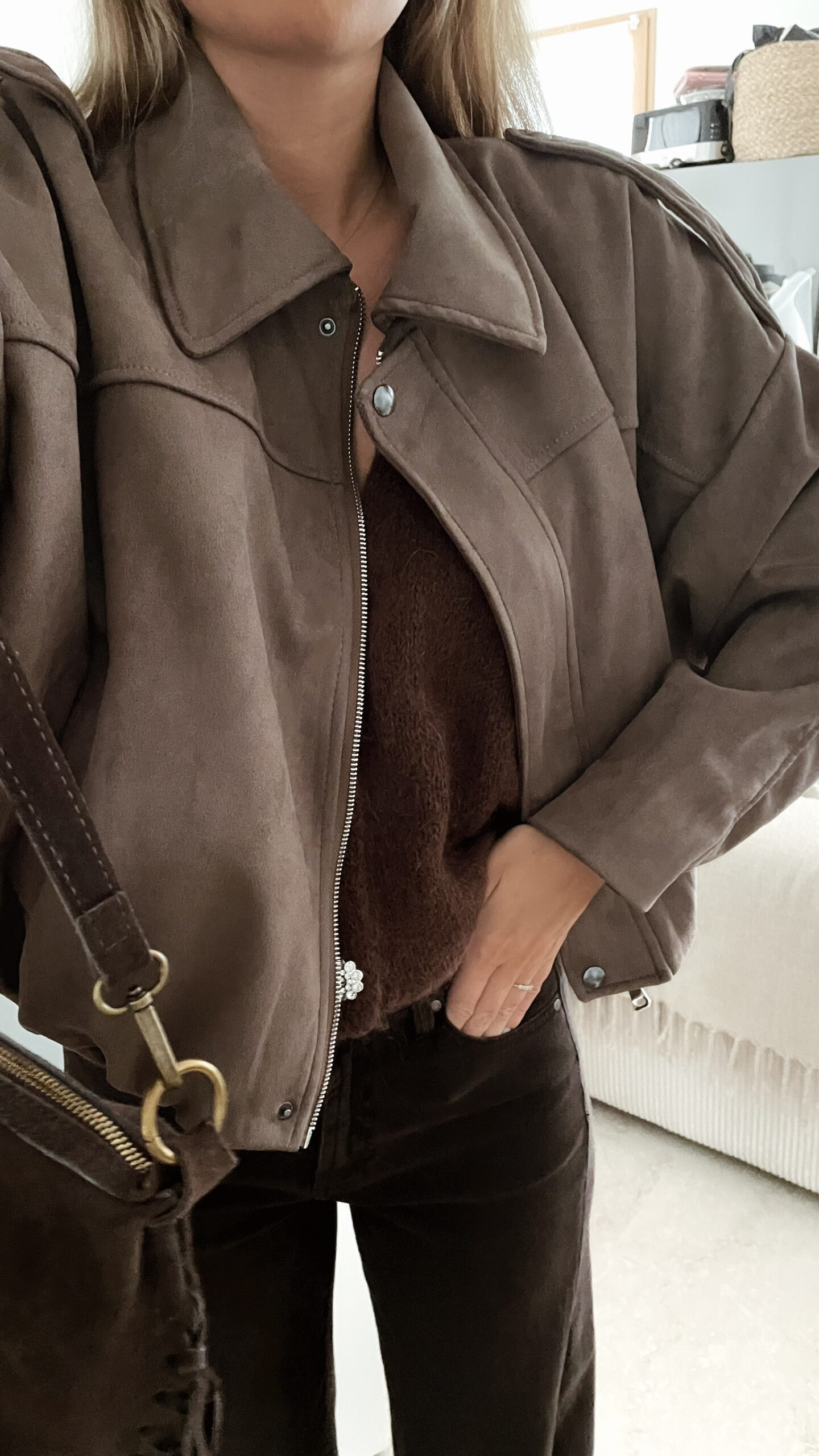 Femme portant une veste effet daim couleur marron taupe, assortie à un pull en alpaga taupe ton sur ton, un pantalon marron, et un sac en daim marron avec des franges dans un intérieur lumineux.