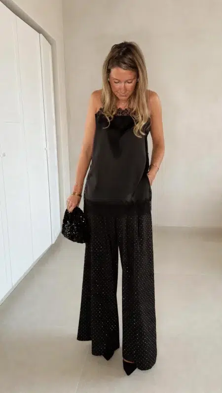Femme portant un top en satin noir avec dentelle et un pantalon large noir à strass, accompagné d’une pochette brillante.