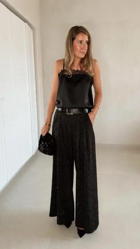 Femme portant un top en satin noir avec dentelle et un pantalon large noir à strass, accompagné d’une pochette brillante.