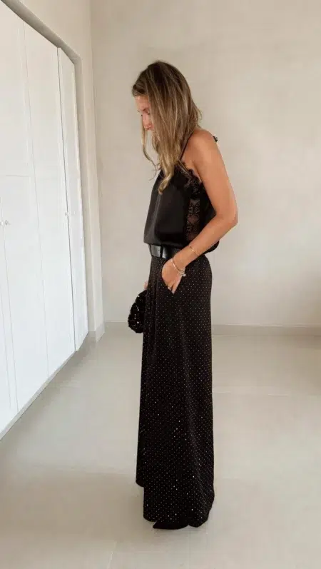 Femme portant un top en satin noir avec dentelle et un pantalon large noir à strass, accompagné d’une pochette brillante.