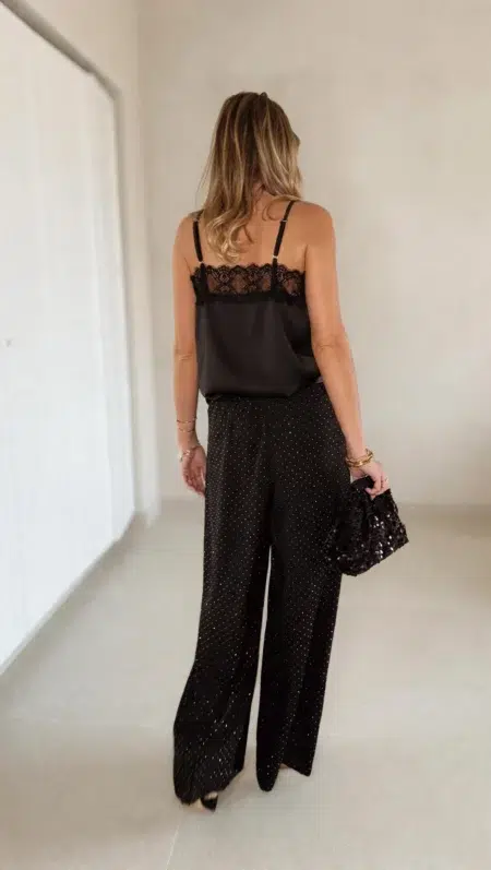 Femme portant un top en satin noir avec dentelle et un pantalon large noir à strass, accompagné d’une pochette brillante.