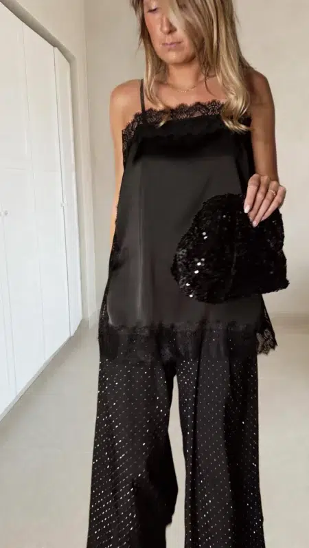 Femme portant un top en satin noir avec dentelle et un pantalon large noir à strass, accompagné d’une pochette brillante.