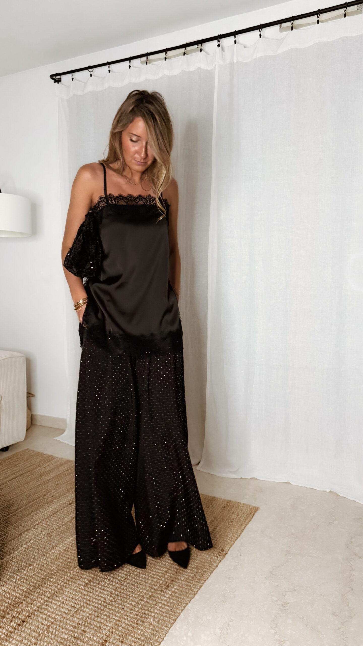 Femme portant un top en satin noir avec dentelle et un pantalon large noir à strass, accompagné d’une pochette brillante.