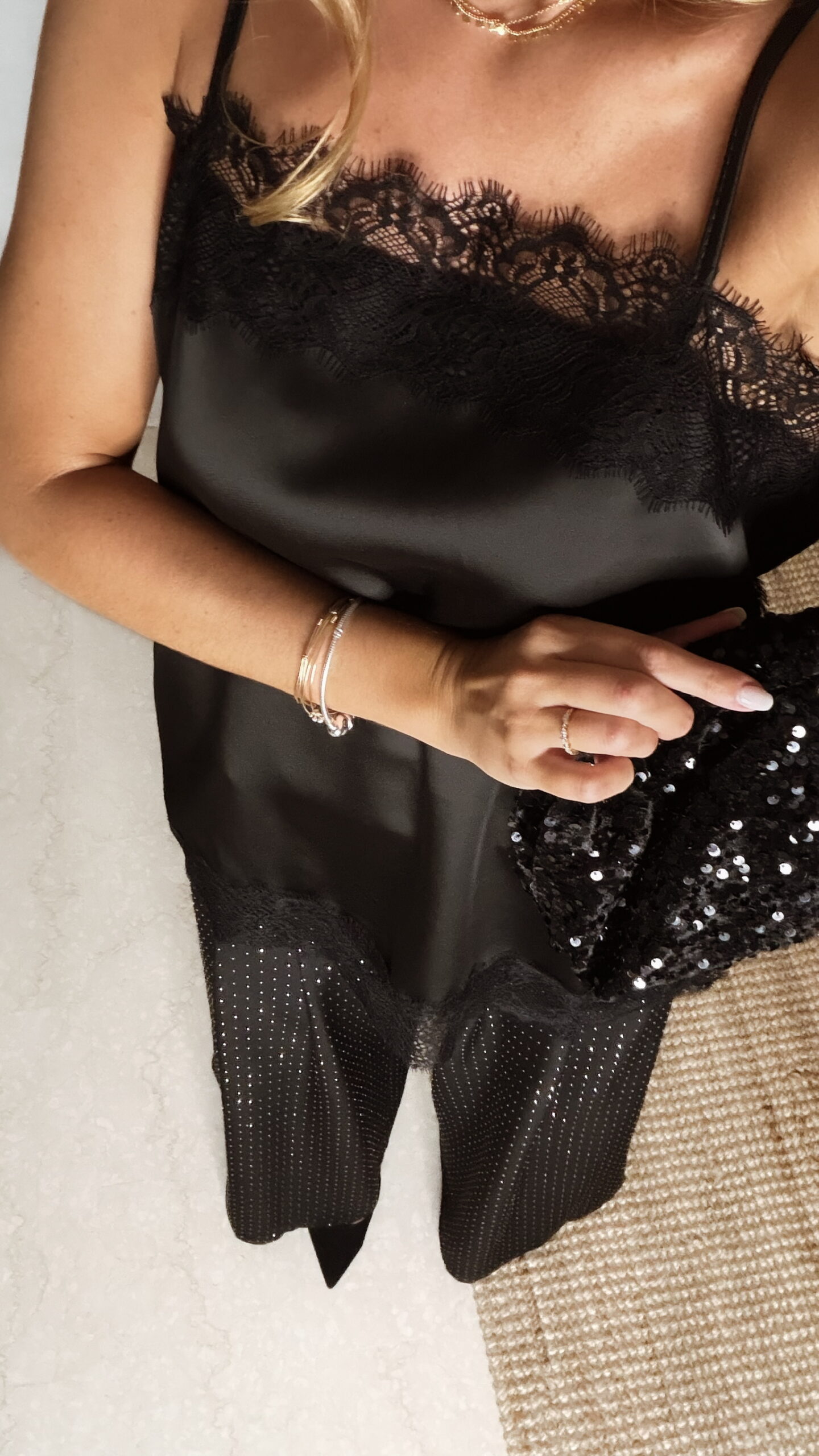 Femme portant un top en satin noir avec dentelle et un pantalon large noir à strass, accompagné d’une pochette brillante.