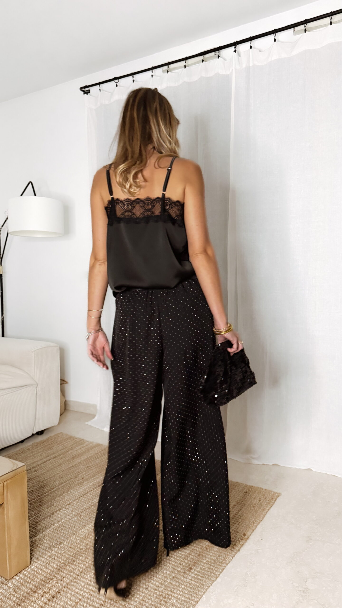 Femme portant un top en satin noir avec dentelle et un pantalon large noir à strass, accompagné d’une pochette brillante.