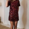 Femme portant une robe courte à sequins bordeaux, coupe ajustée et manches courtes, assortie de chaussures à talons noirs.