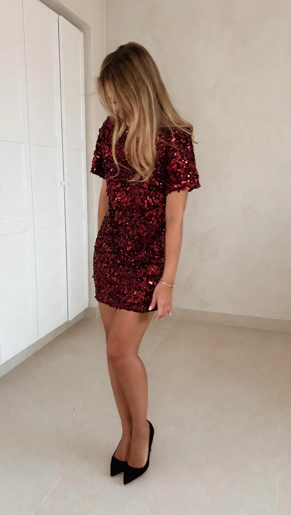 Femme portant une robe courte à sequins bordeaux, coupe ajustée et manches courtes, assortie de chaussures à talons noirs.