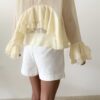Blouse femme jaune pastel en tissu léger avec volants, manches évasées et col à nouer, portée avec un short blanc.