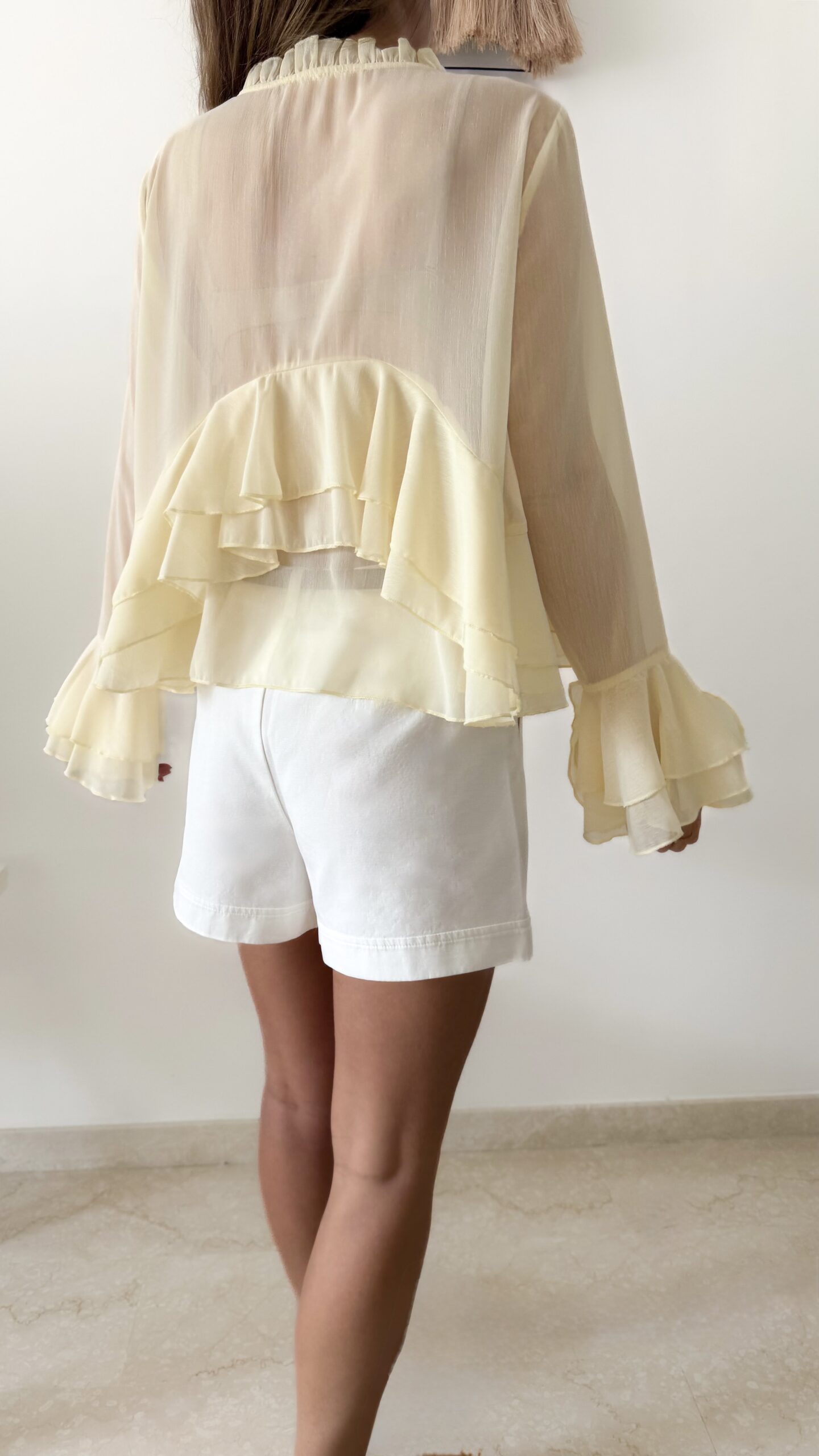 Blouse femme jaune pastel en tissu léger avec volants, manches évasées et col à nouer, portée avec un short blanc.
