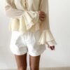 Blouse femme jaune pastel en tissu léger avec volants, manches évasées et col à nouer, portée avec un short blanc.