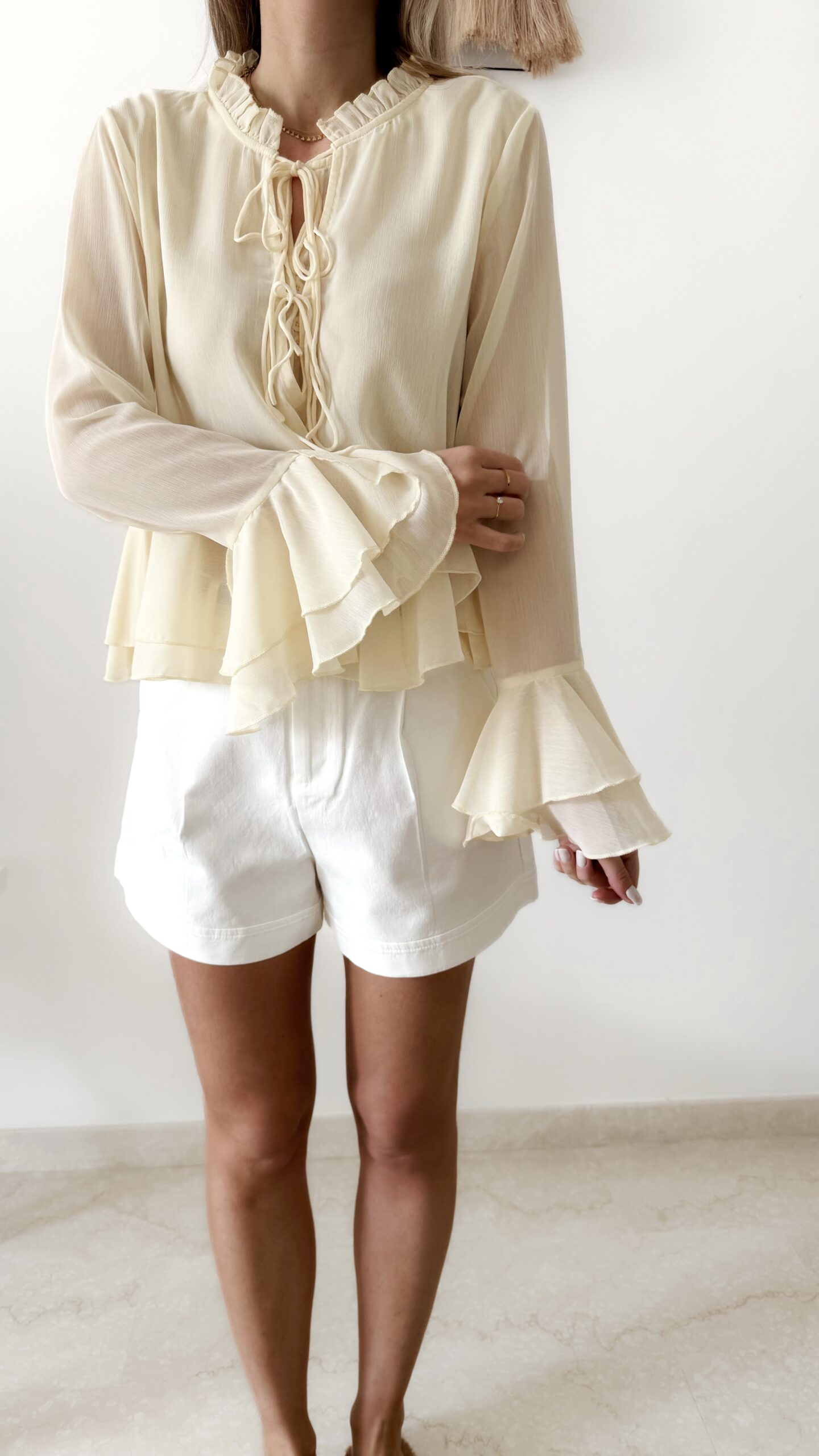 Blouse femme jaune pastel en tissu léger avec volants, manches évasées et col à nouer, portée avec un short blanc.