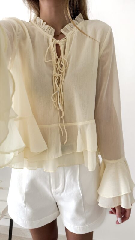 Blouse femme jaune pastel en tissu léger avec volants, manches évasées et col à nouer, portée avec un short blanc.