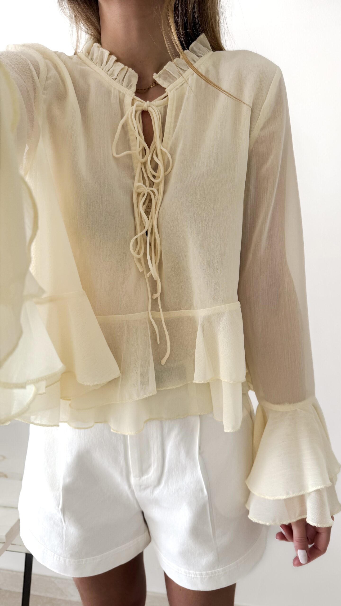 Blouse femme jaune pastel en tissu léger avec volants, manches évasées et col à nouer, portée avec un short blanc.