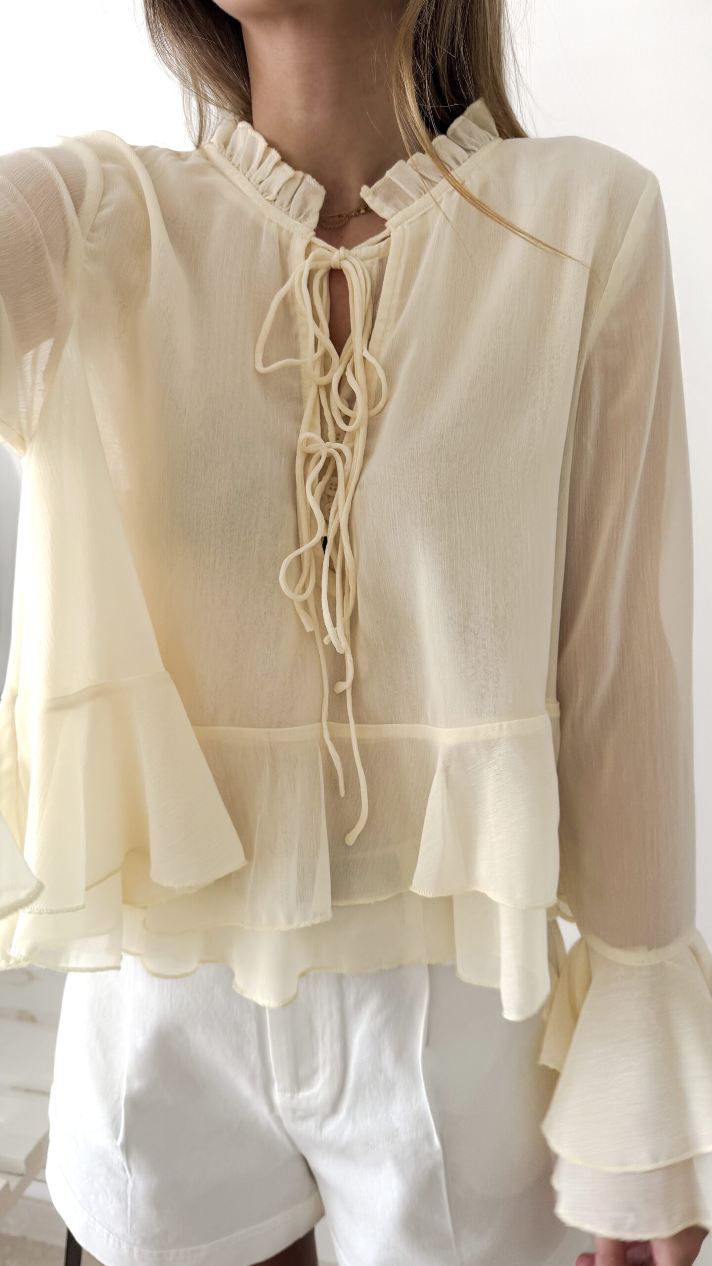 Blouse femme jaune pastel en tissu léger avec volants, manches évasées et col à nouer, portée avec un short blanc.