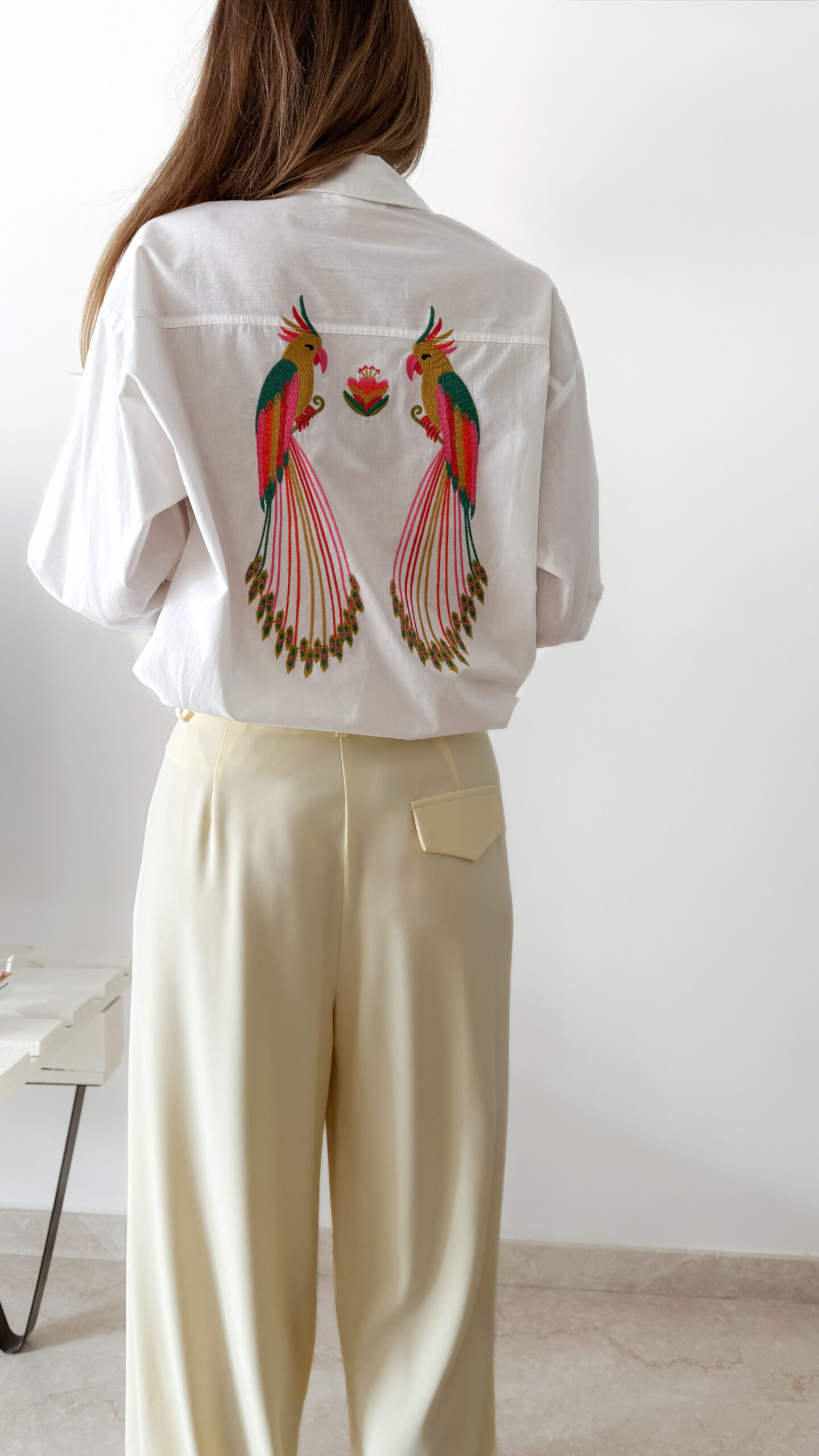 Chemise blanche femme avec broderie de deux perroquets colorés dans le dos, portée avec un pantalon jaune pâle