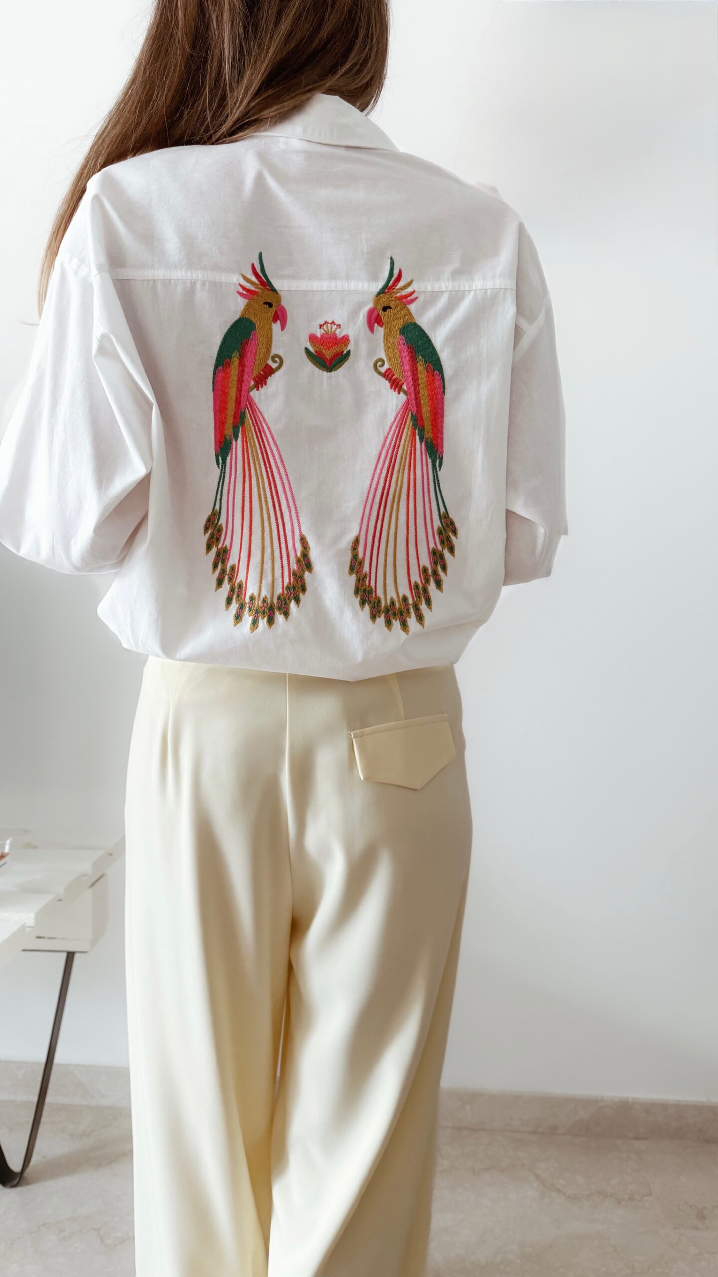 Chemise blanche femme avec broderie de deux perroquets colorés dans le dos, portée avec un pantalon jaune pâle