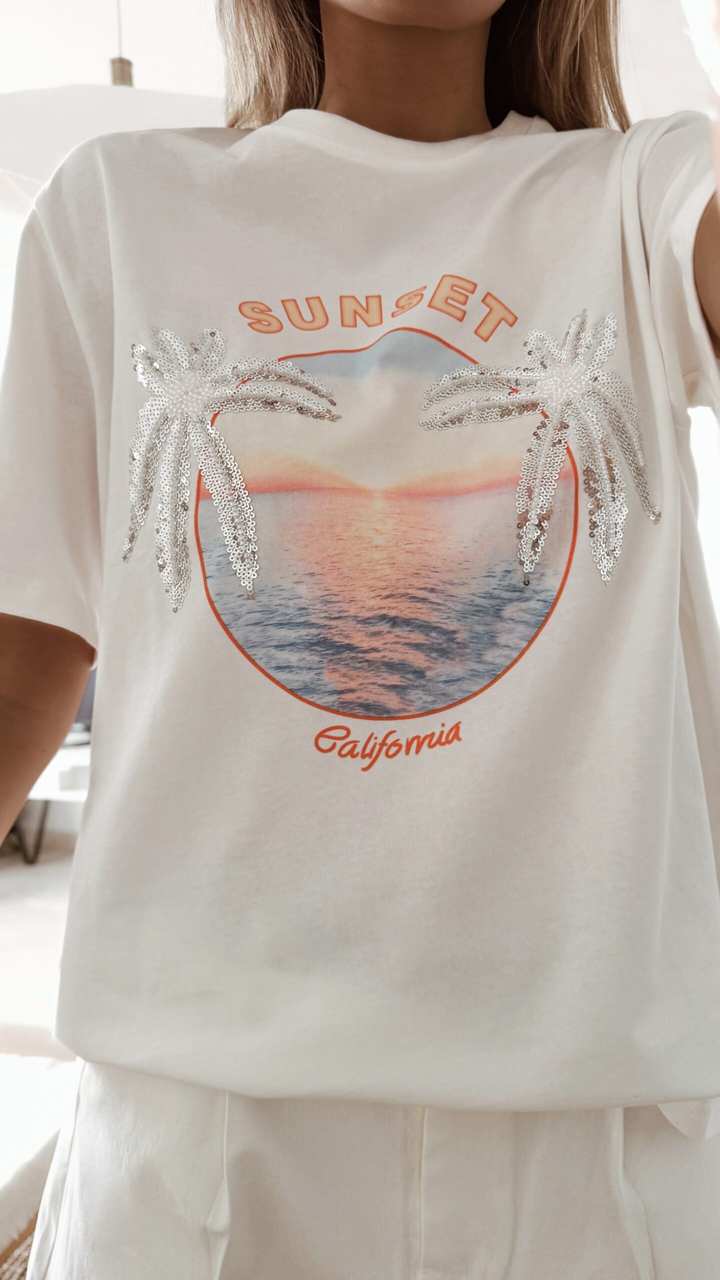 Femme portant un t-shirt blanc avec imprimé coucher de soleil et palmiers en sequins