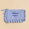 Collection de pochettes colorées à rayures et motifs vichy avec volants décoratifs et messages brodés, présentées sur un fond beige dans un style féminin et tendance.Rayures, vichy, volants et messages brodés : les accessoires parfaits pour glisser un peu de bonne humeur et beaucoup de style dans votre quotidien. À emporter partout, de la plage à la ville. ✨