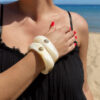 Femme portant deux bracelets rigides nacrés écrus avec détails dorés en forme de soleil sur une plage ensoleillée, look élégant et estival avec robe noire fluide.