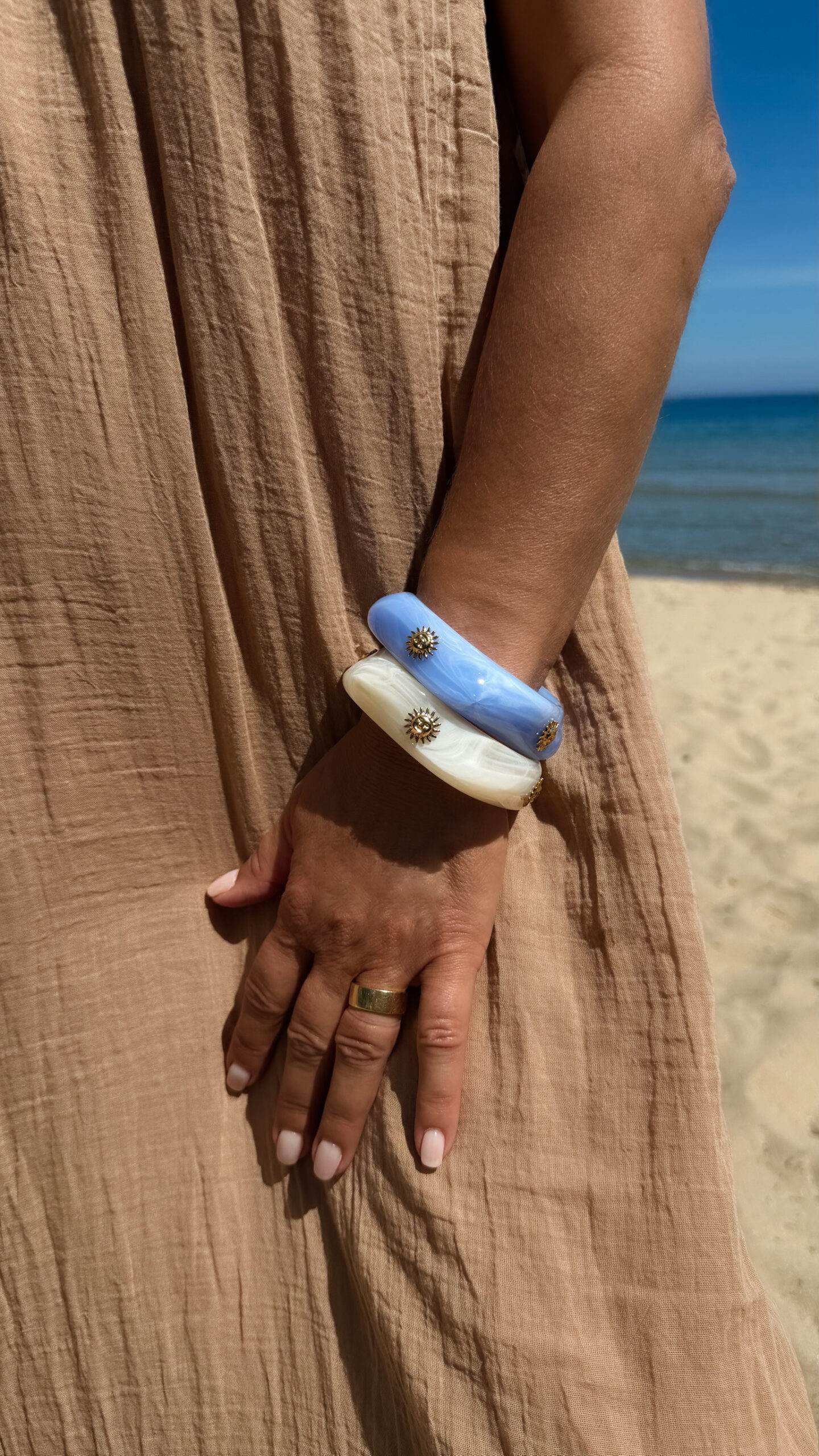 Femme portant deux bracelets rigides nacrés écrus avec détails dorés en forme de soleil sur une plage ensoleillée, look élégant et estival avec robe noire fluide.