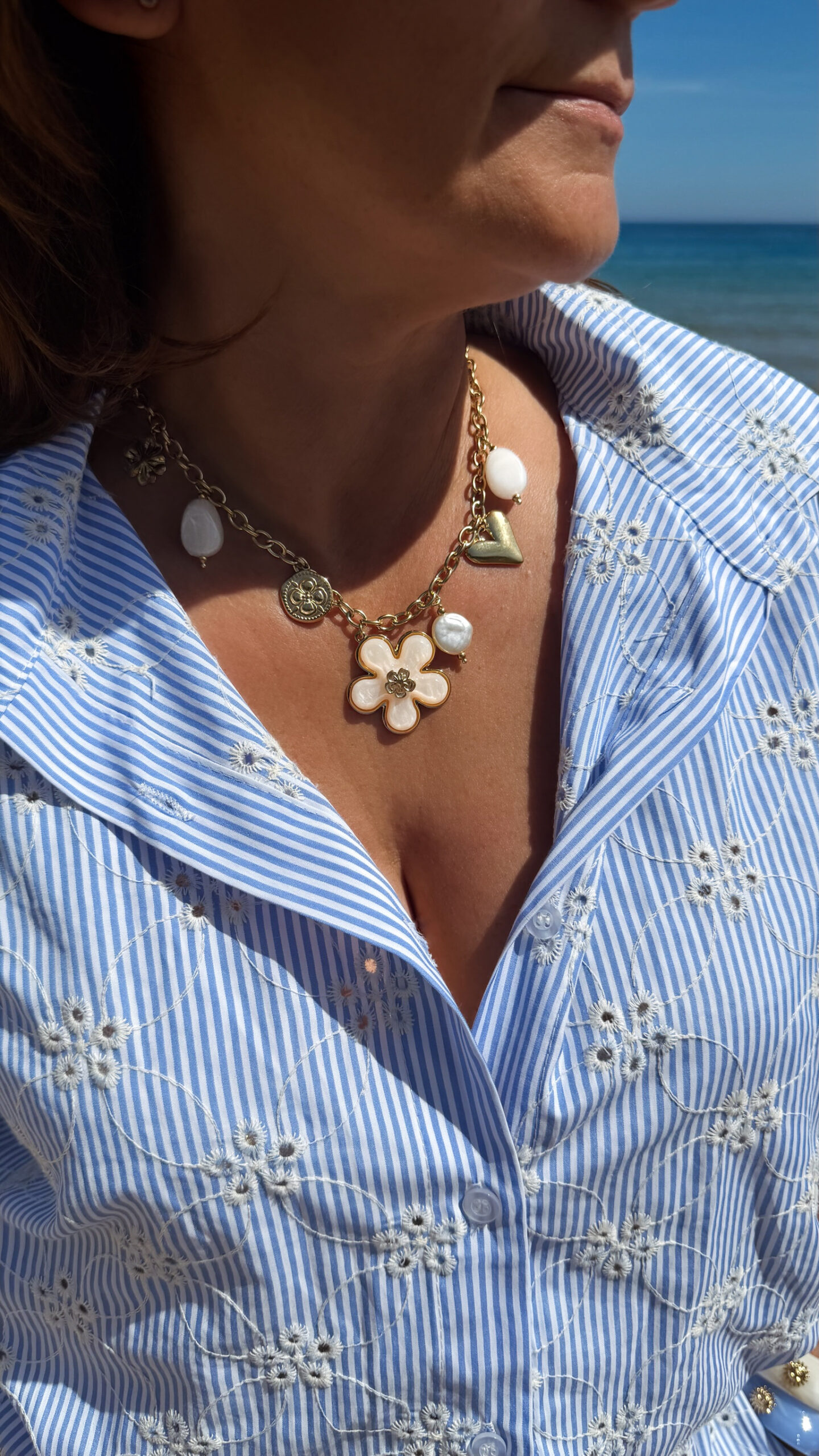 Collier chaîne dorée avec pendentif fleur blanche, perle et charms dorés porté sur peau bronzée avec tenue estivale.