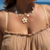 Collier chaîne dorée avec pendentif fleur blanche, perle et charms dorés porté sur peau bronzée avec tenue estivale.