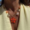 Collier chaîne dorée avec pendentif fleur corail, perle et charms colorés porté sur peau bronzée avec tenue estivale.