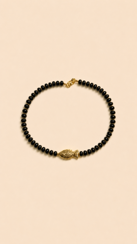 Collier en perles noires avec pendentif poisson doré porté autour du cou sur peau bronzée, look estival élégant.