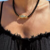 Collier en perles noires avec pendentif poisson doré porté autour du cou sur peau bronzée, look estival élégant.