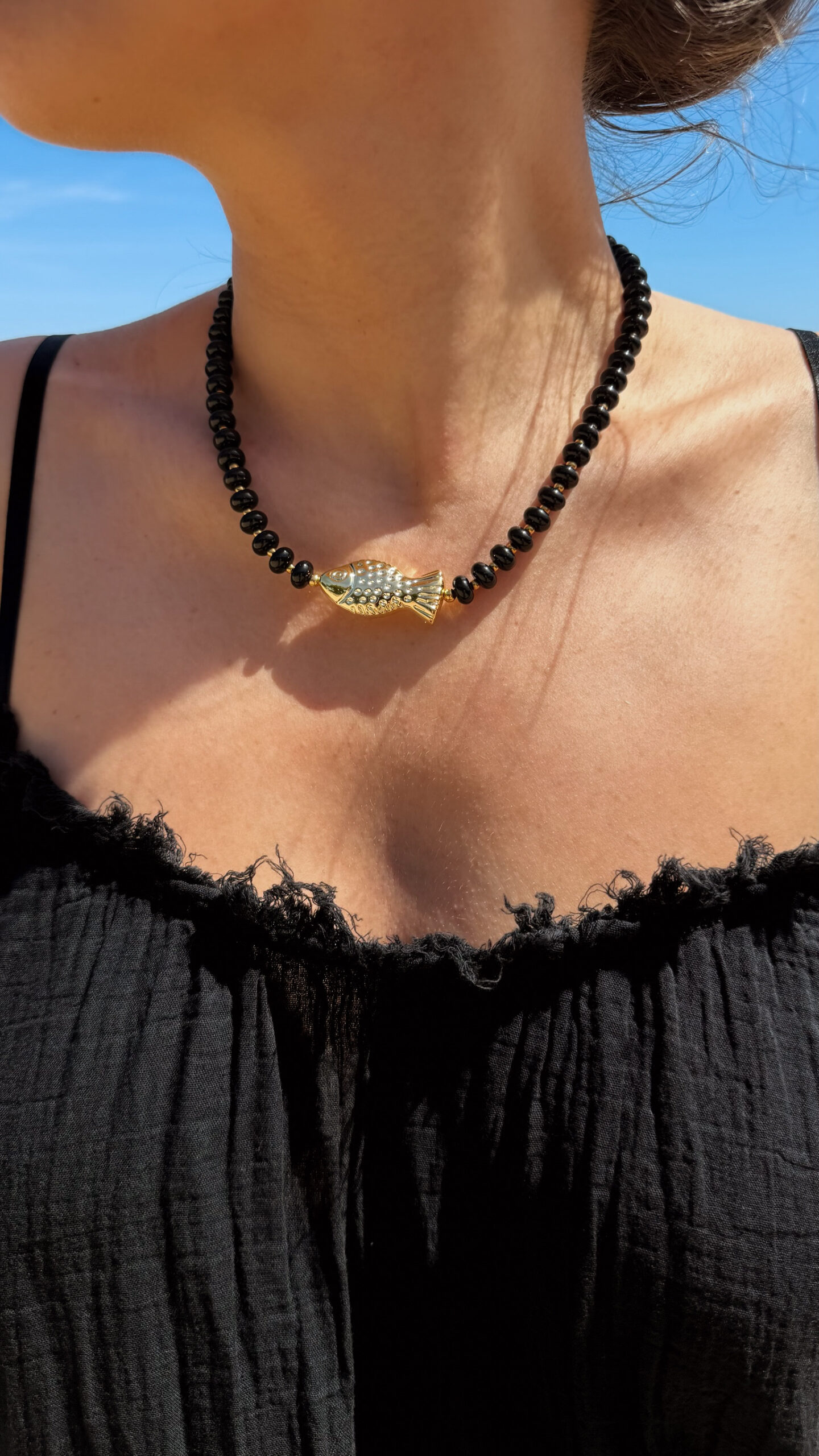 Collier en perles noires avec pendentif poisson doré porté autour du cou sur peau bronzée, look estival élégant.
