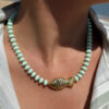 Collier en perles vert amande avec pendentif poisson doré porté autour du cou sur peau bronzée, look estival élégant.