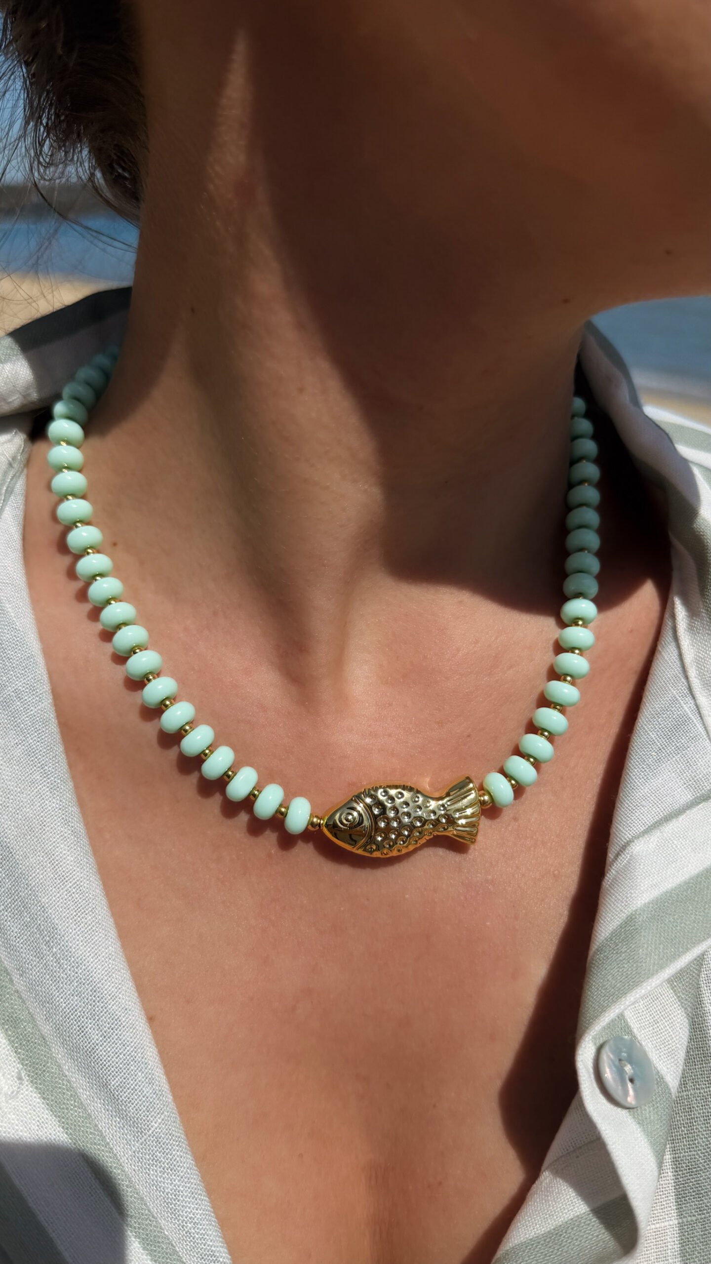 Collier en perles vert amande avec pendentif poisson doré porté autour du cou sur peau bronzée, look estival élégant.