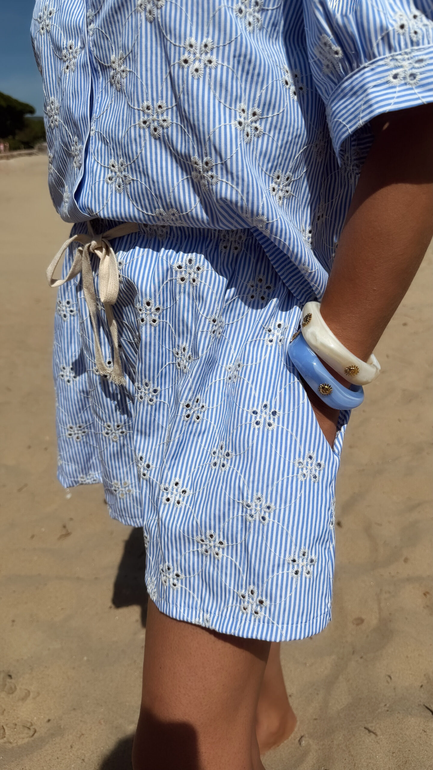 Femme portant deux bracelets rigides nacrés vert avec détails dorés en forme de soleil sur une plage ensoleillée, look élégant et estival