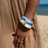 Femme portant deux bracelets rigides nacrés vert avec détails dorés en forme de soleil sur une plage ensoleillée, look élégant et estival