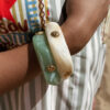 Femme portant deux bracelets rigides nacrés vert avec détails dorés en forme de soleil sur une plage ensoleillée, look élégant et estival