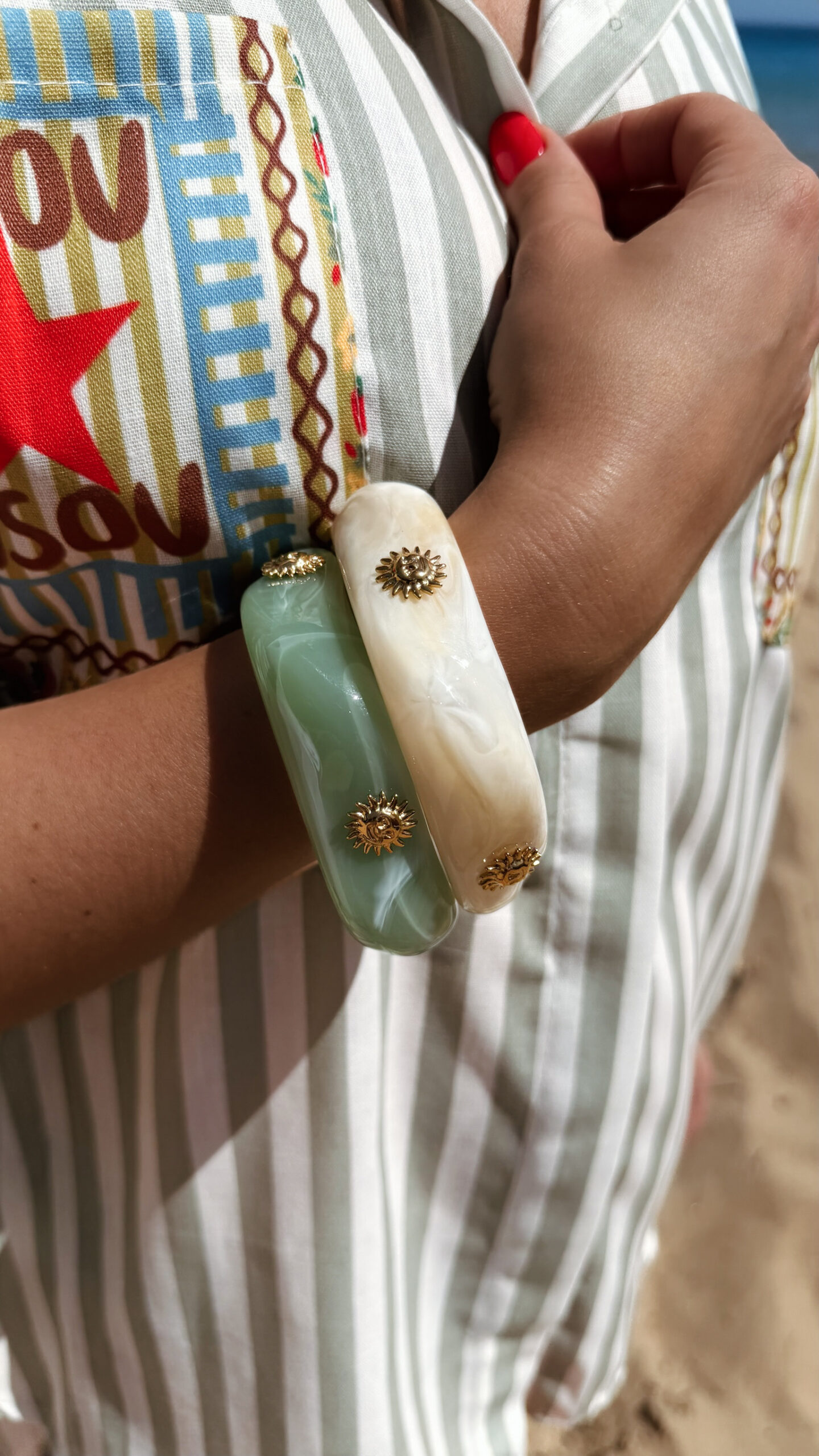 Femme portant deux bracelets rigides nacrés vert avec détails dorés en forme de soleil sur une plage ensoleillée, look élégant et estival