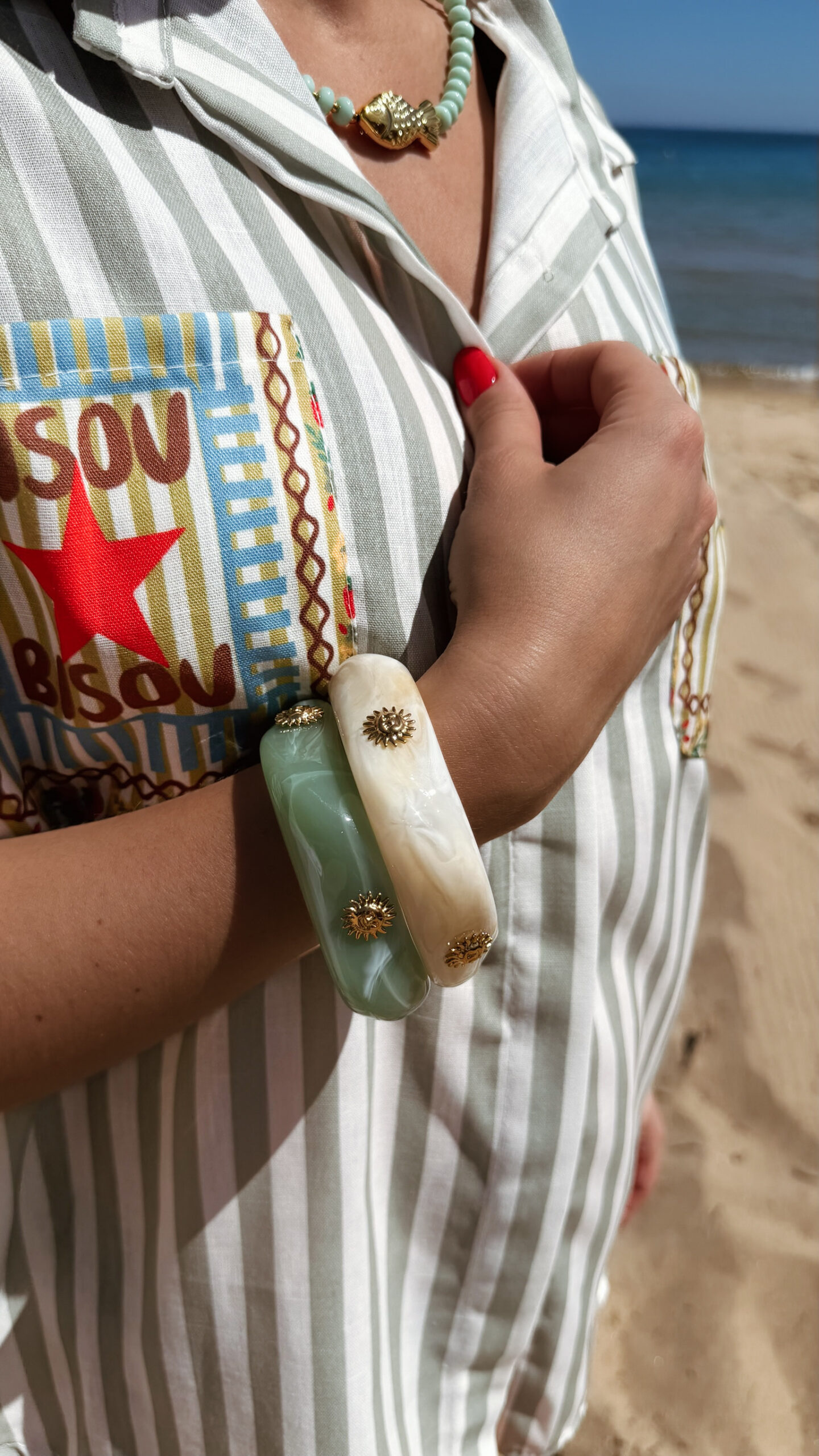 Femme portant deux bracelets rigides nacrés vert avec détails dorés en forme de soleil sur une plage ensoleillée, look élégant et estival