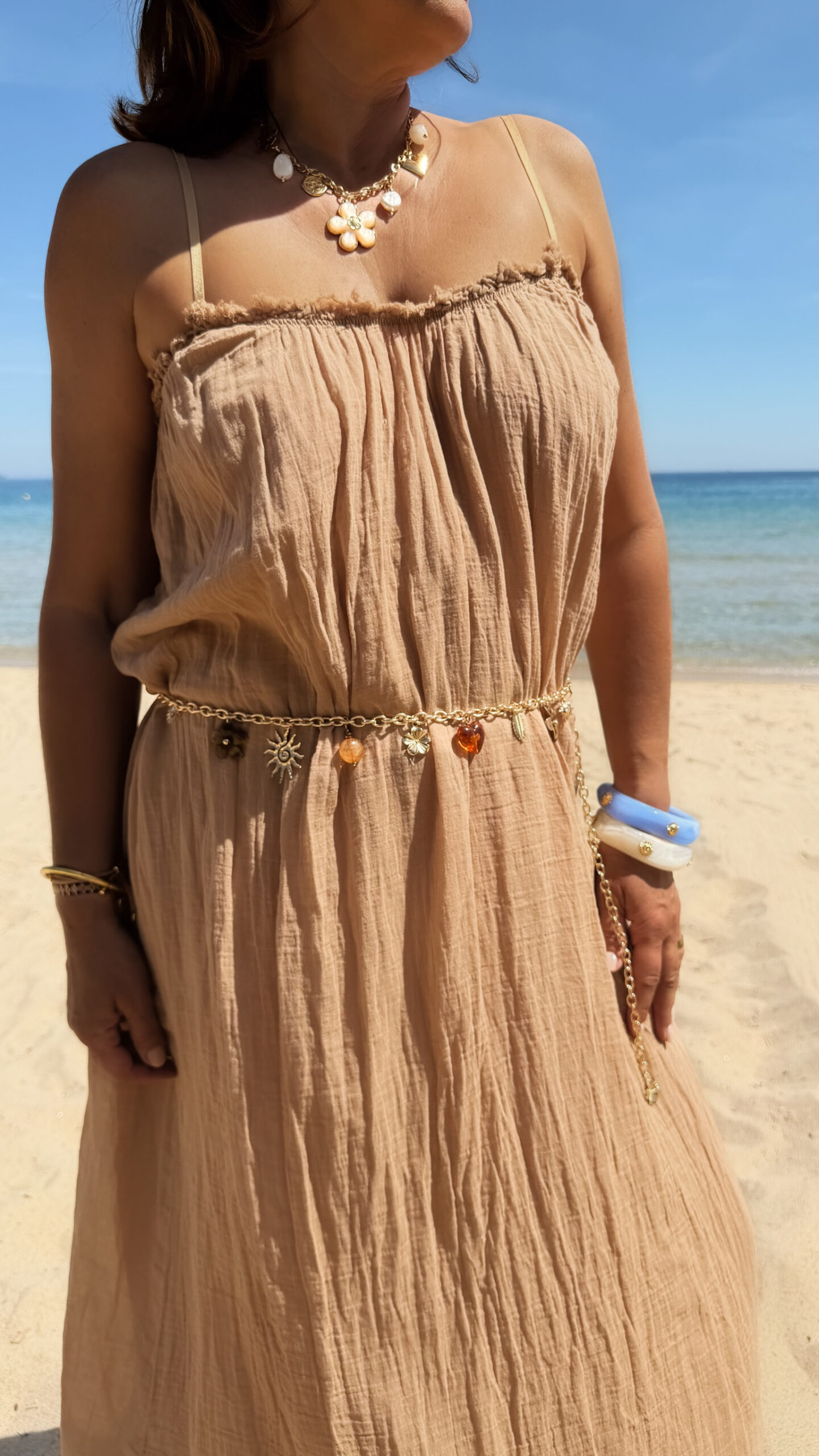 Ceinture chaîne dorée ornée de fleurs marrons et breloques décoratives portée sur une robe fluide foncée en bord de mer.