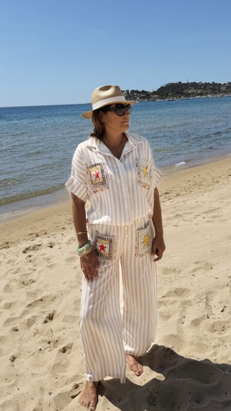 Femme portant l'ensemble Zoé composé d'une blouse rayée beige brodée et d'un pantalon fluide rayé beige sur une plage ensoleillée.