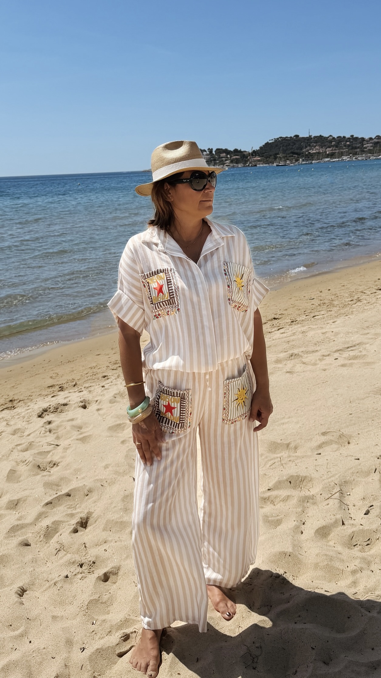 Femme portant l'ensemble Zoé composé d'une blouse rayée beige brodée et d'un pantalon fluide rayé beige sur une plage ensoleillée.