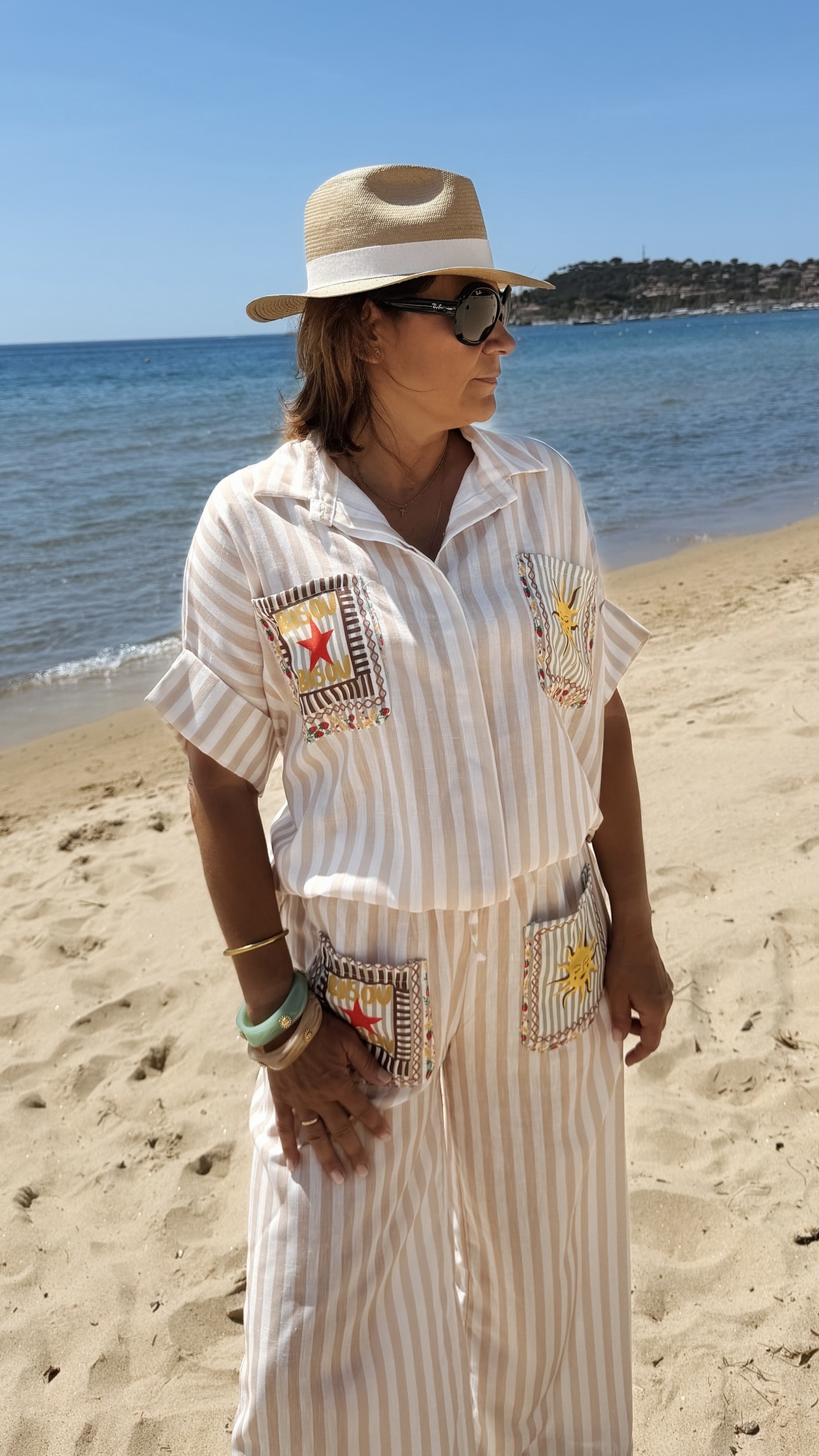 Femme portant l'ensemble Zoé composé d'une blouse rayée beige brodée et d'un pantalon fluide rayé beige sur une plage ensoleillée.
