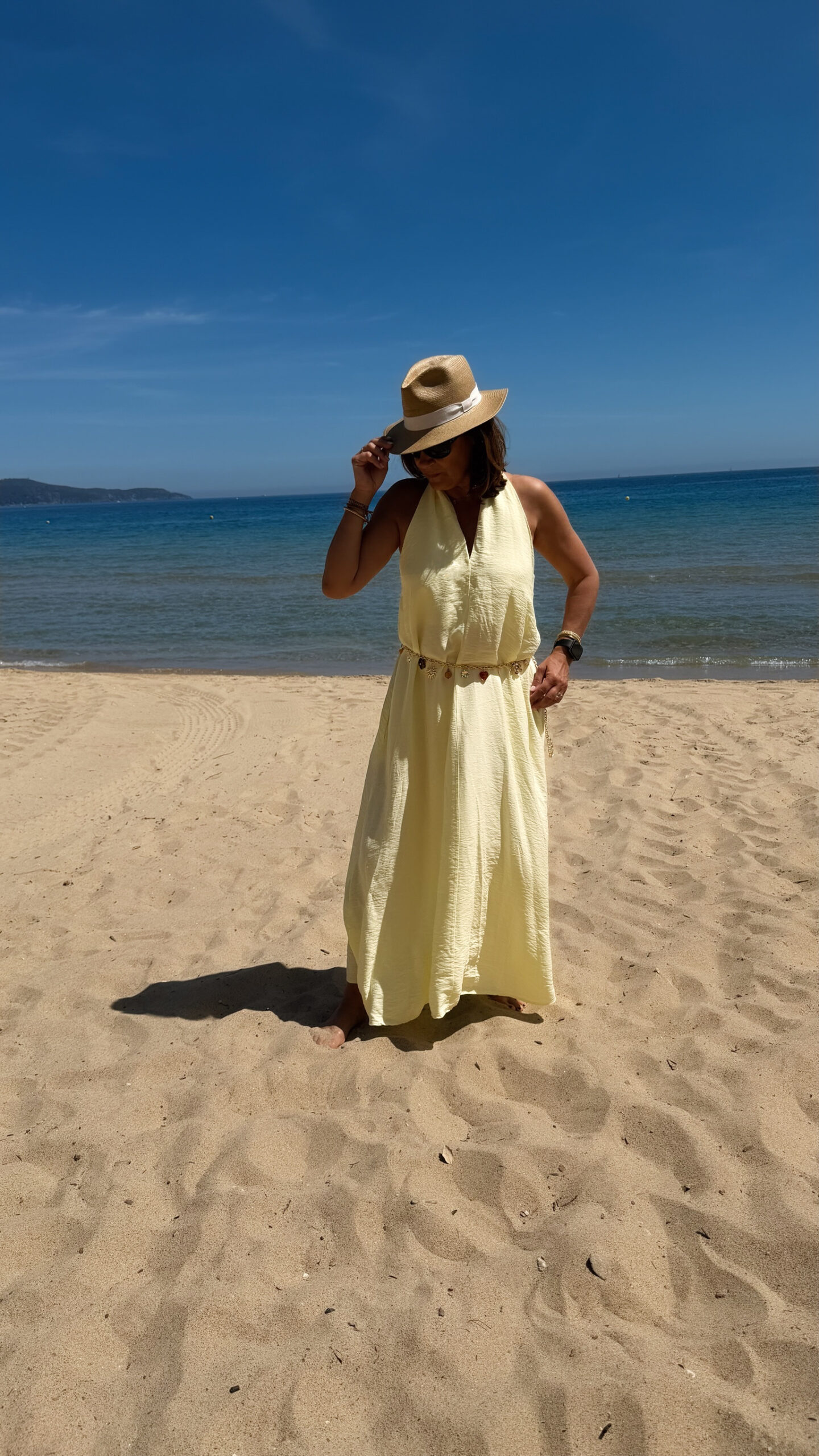 Robe longue jaune dos nu Carla pour femme, coupe fluide et élégante, portée sur une plage en bord de mer sous le soleil d'été.
