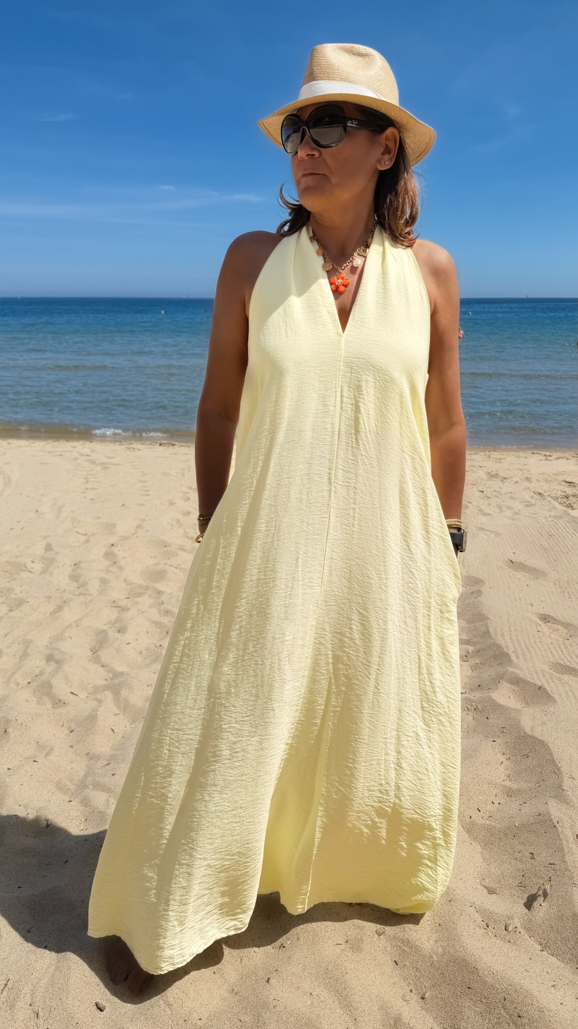 Robe longue jaune dos nu Carla pour femme, coupe fluide et élégante, portée sur une plage en bord de mer sous le soleil d'été.