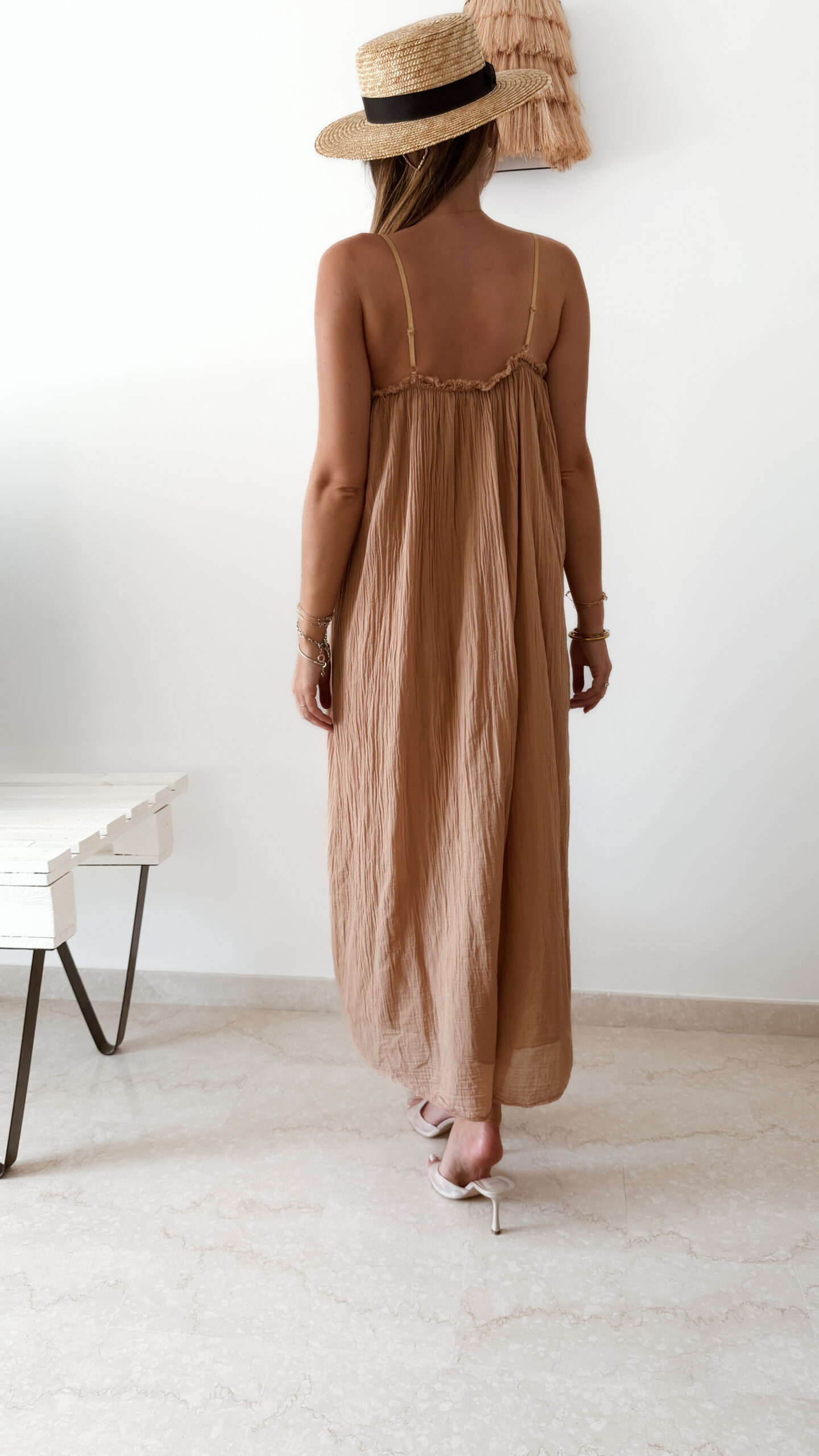 Femme portant une robe longue camel en gaze de coton sur une plage ensoleillée, avec chapeau et lunettes de soleil.